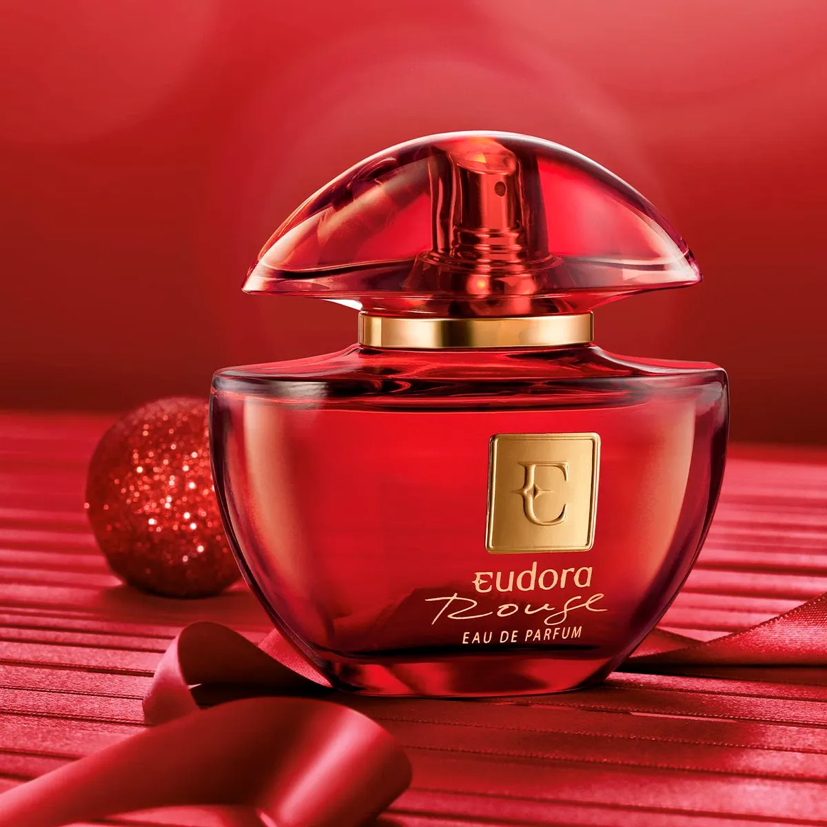 Frasco do perfume Eudora Rouge sobre superfície escura com fundo elegante, iluminado suavemente para destacar seu tom vermelho profundo e detalhes dourados.