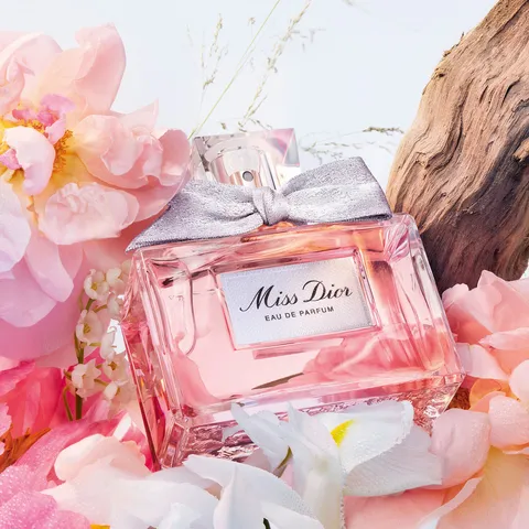 Frasco do perfume Miss Dior Eau de Parfum com laço rosa, rodeado por flores delicadas sobre fundo claro e elegante.