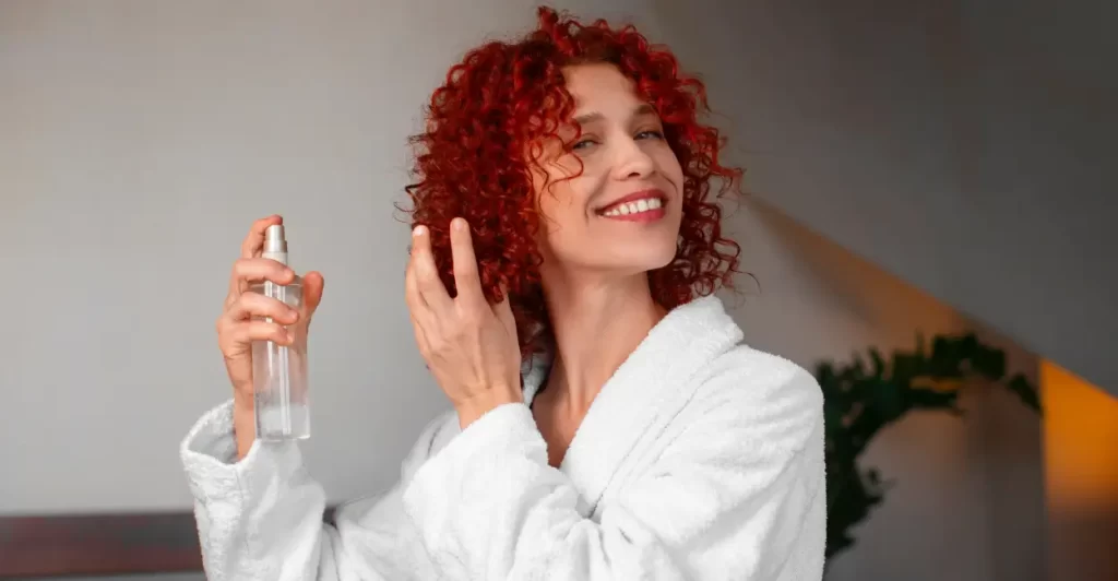 Mulher borrifando perfume para cabelo nos fios cacheados e ruivos, mostrando o uso do produto na rotina de beleza.