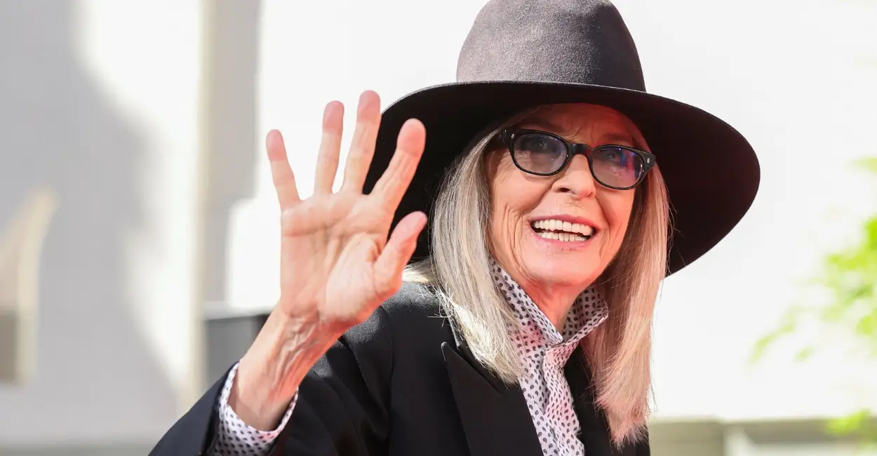 Diane Keaton sorrindo e acenando, com chapéu preto e óculos escuros