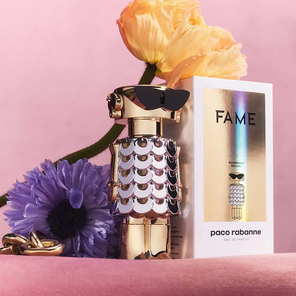 Frasco dourado do perfume Fame da Paco Rabanne em formato de robô estilizado, com flores e embalagem ao fundo sobre fundo rosa.