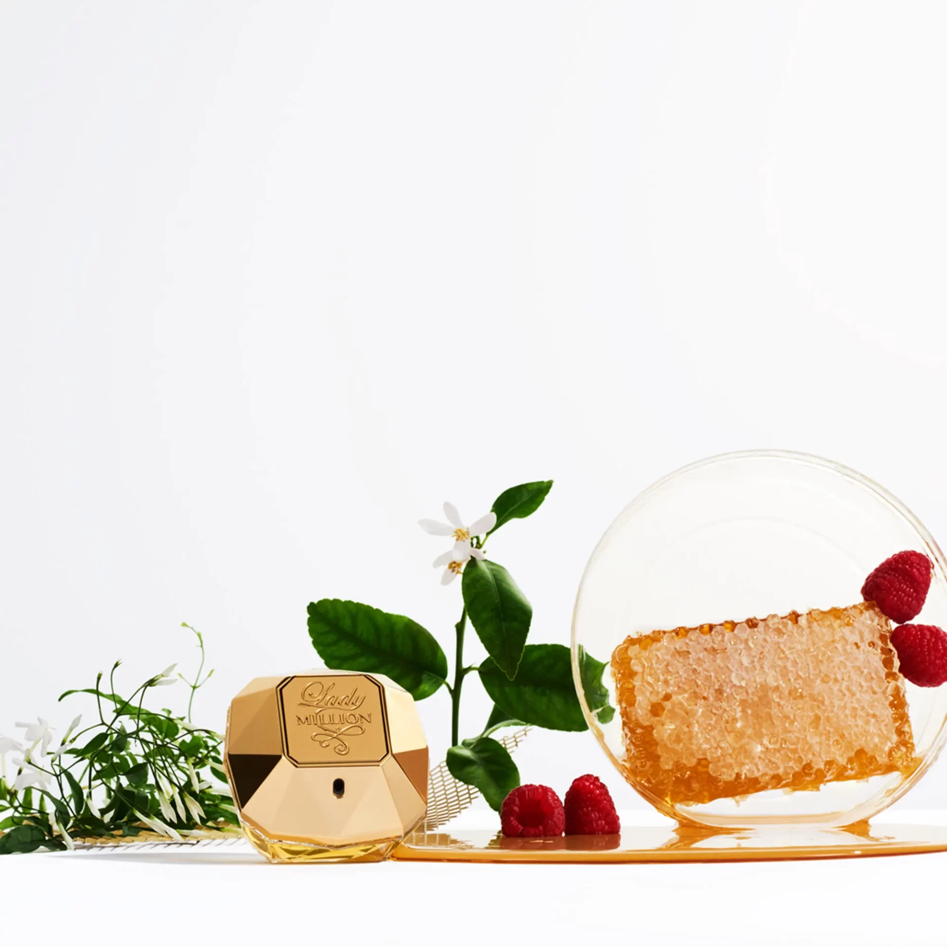 Frasco dourado do perfume Lady Million ao lado de framboesas, favo de mel, flores brancas e folhas verdes sobre fundo branco.
