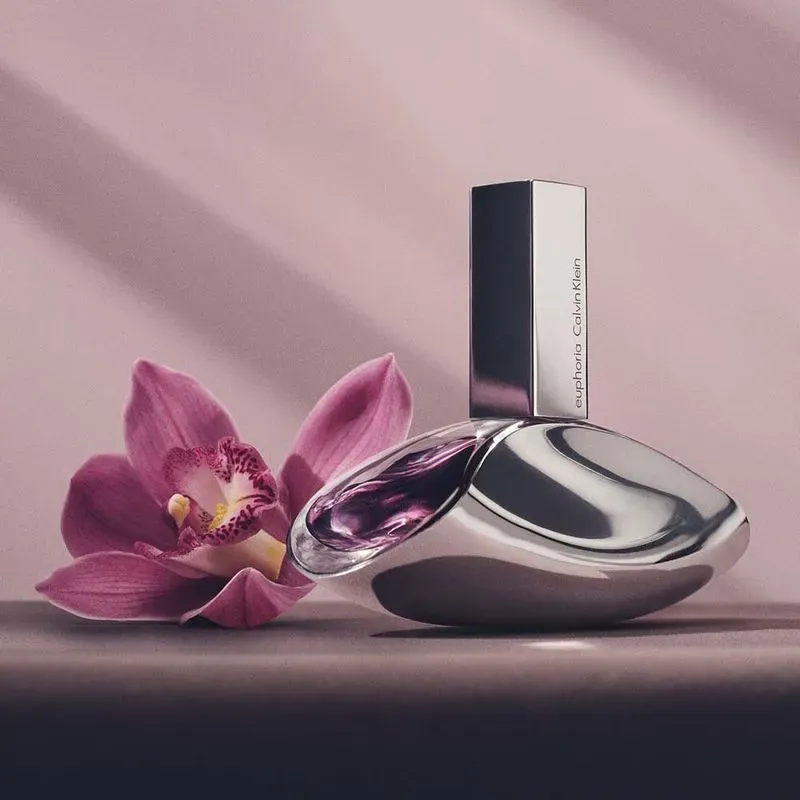 Frasco prateado do Euphoria Calvin Klein Eau de Parfum ao lado de uma orquídea rosa sobre fundo rosé, iluminação suave.